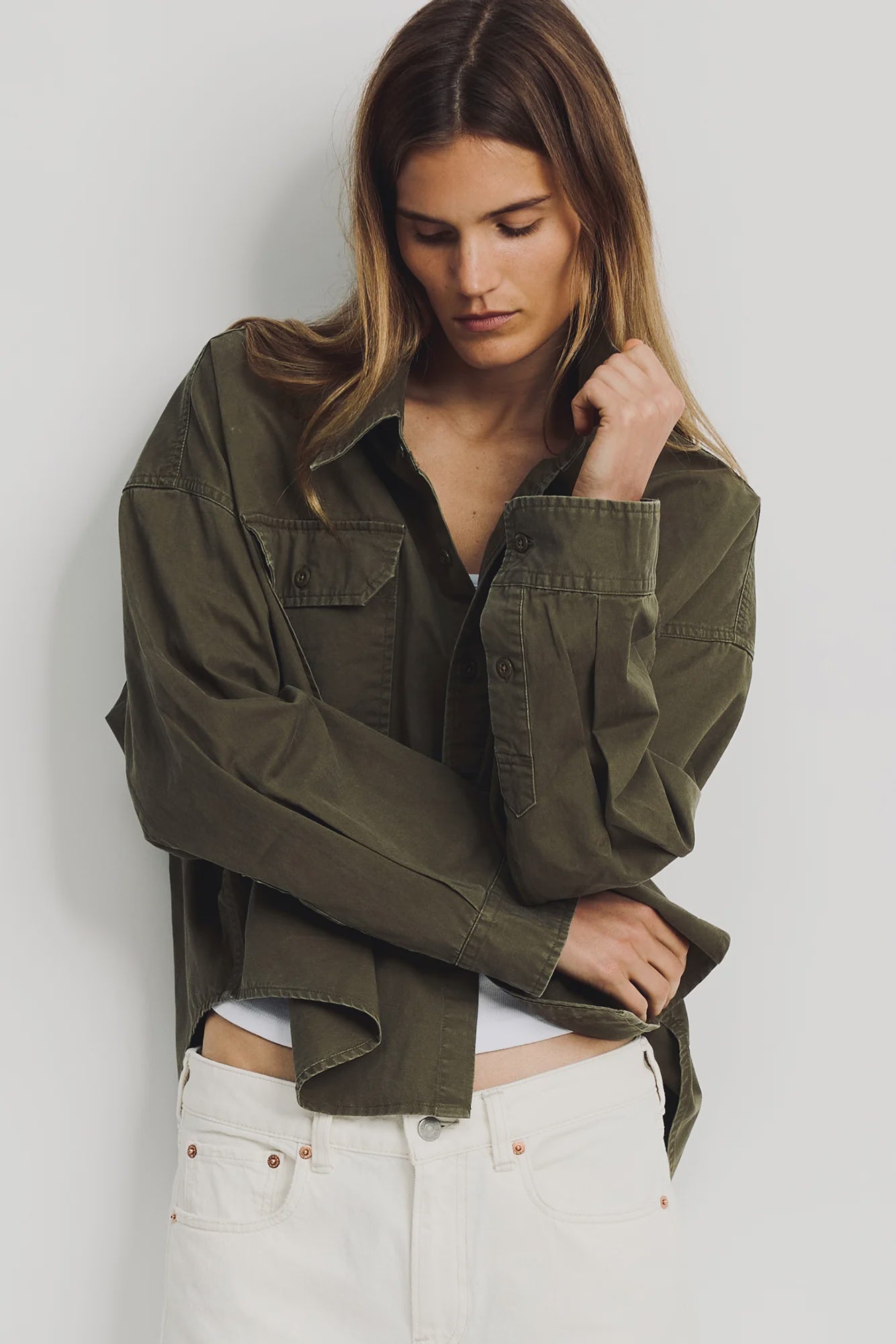 A woman with long brown hair looks down, wearing the Denimist CROPPED UTILITY SHIRT OLIVE over a white top and white pants, standing against a plain light background.