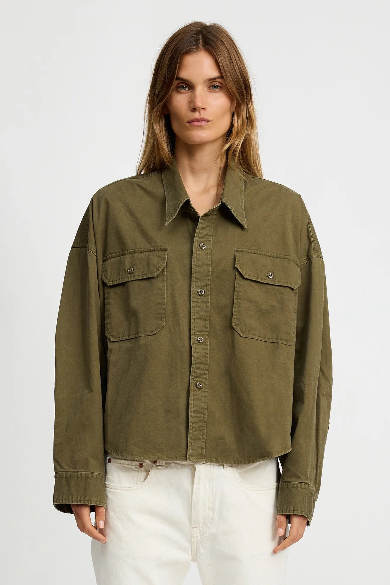 A woman with long, light brown hair wears the Denimist CROPPED UTILITY SHIRT OLIVE, a boxy button-up with flap chest pockets, paired with white pants against a plain light background.