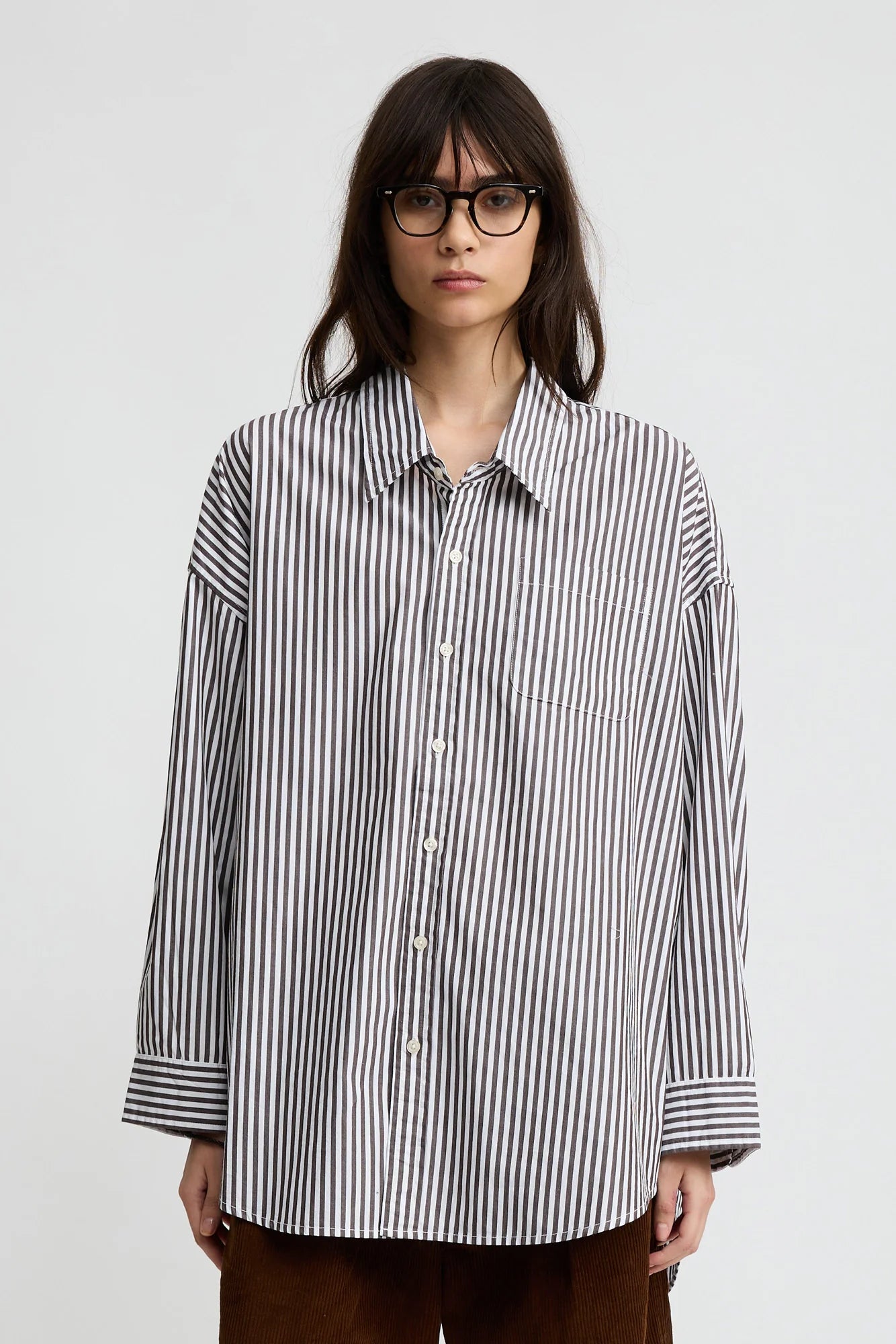 A person with long dark hair and glasses wears a relaxed fit Button Front Shirt by Denimist in black-and-white vertical stripes and brown pants, standing against a plain light gray background.