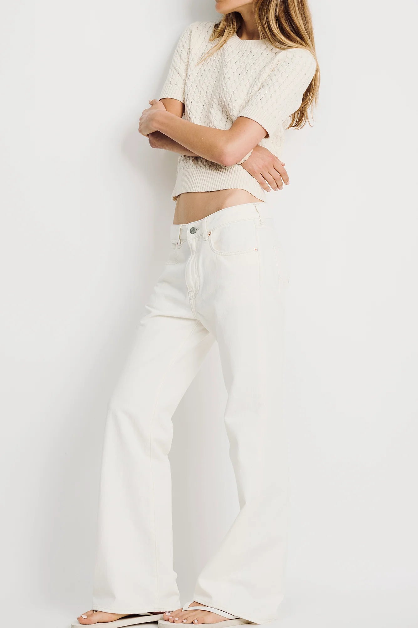 A woman stands barefoot against a white backdrop, arms crossed and face out of frame, wearing Denimist’s SHORT SLEEVE CABLE CREWNECK in beige with high-waisted white jeans.