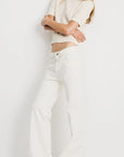 A woman stands barefoot against a white backdrop, arms crossed and face out of frame, wearing Denimist’s SHORT SLEEVE CABLE CREWNECK in beige with high-waisted white jeans.