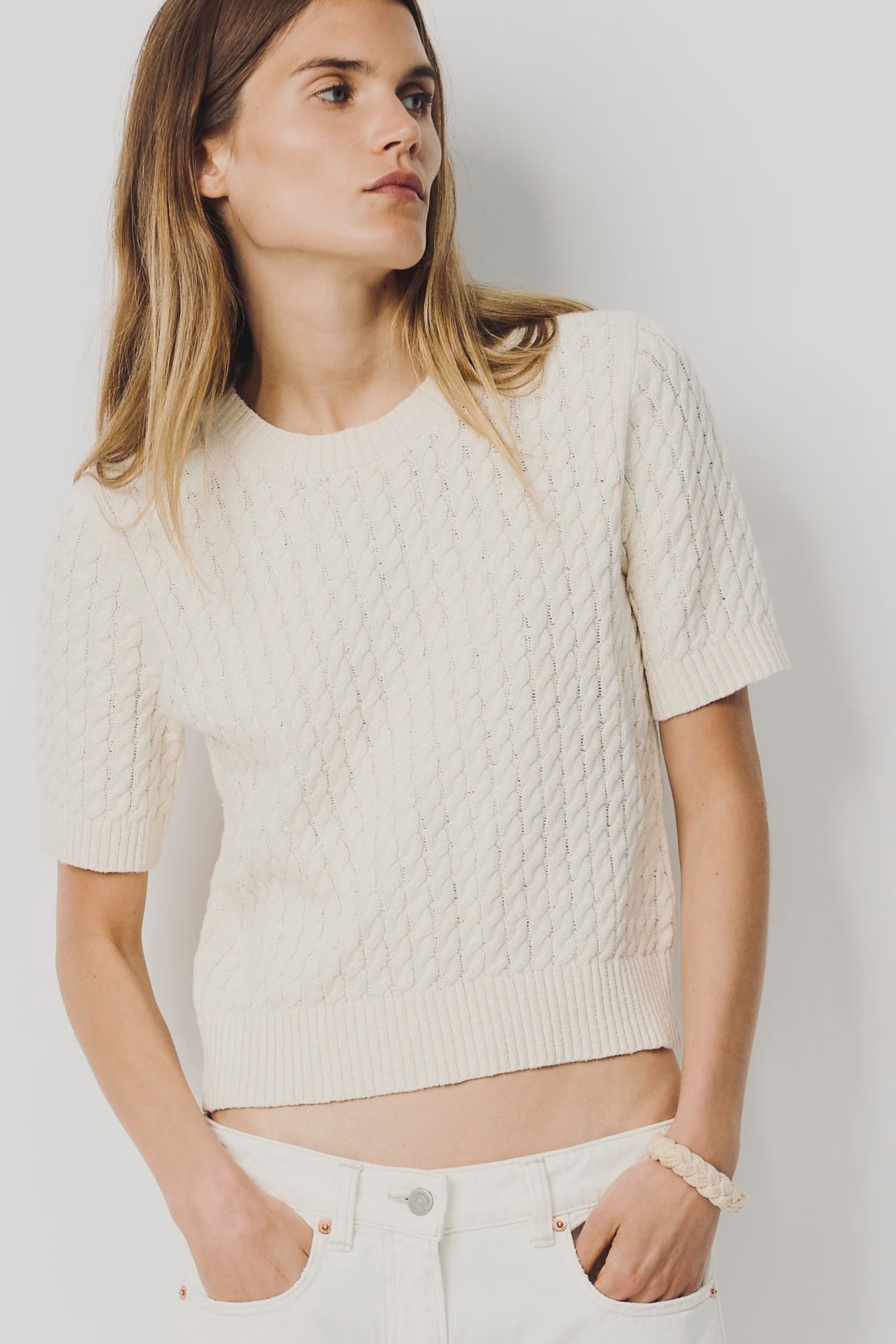 A woman with long, straight hair models the Denimist SHORT SLEEVE CABLE CREWNECK—an off-white, 100% cotton sweater—paired with white pants. She stands against a plain light background, one hand in her pocket.