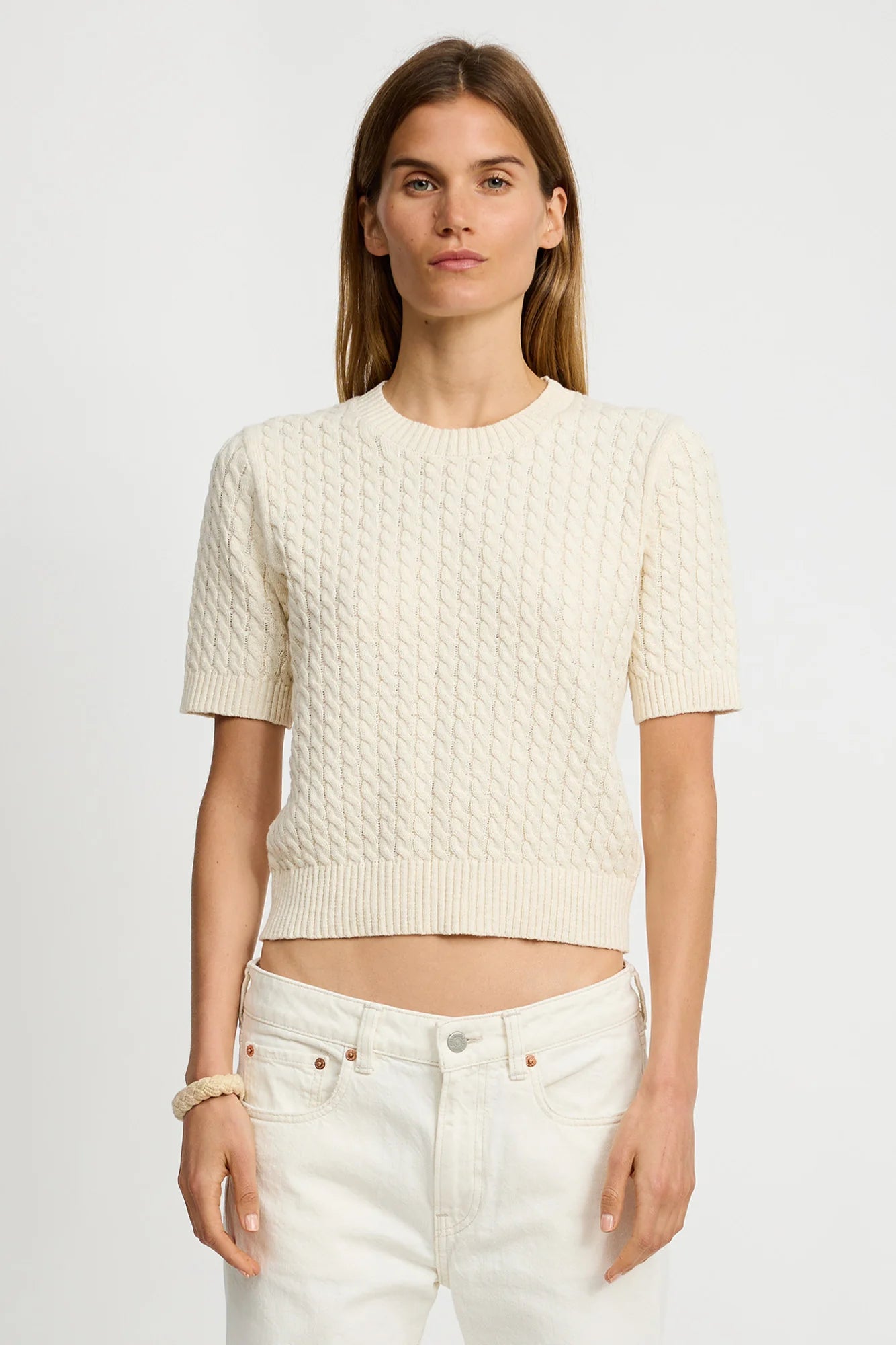 A woman models the Denimist SHORT SLEEVE CABLE CREWNECK in cream, paired with high-waisted white jeans, standing against a plain white background and looking at the camera.