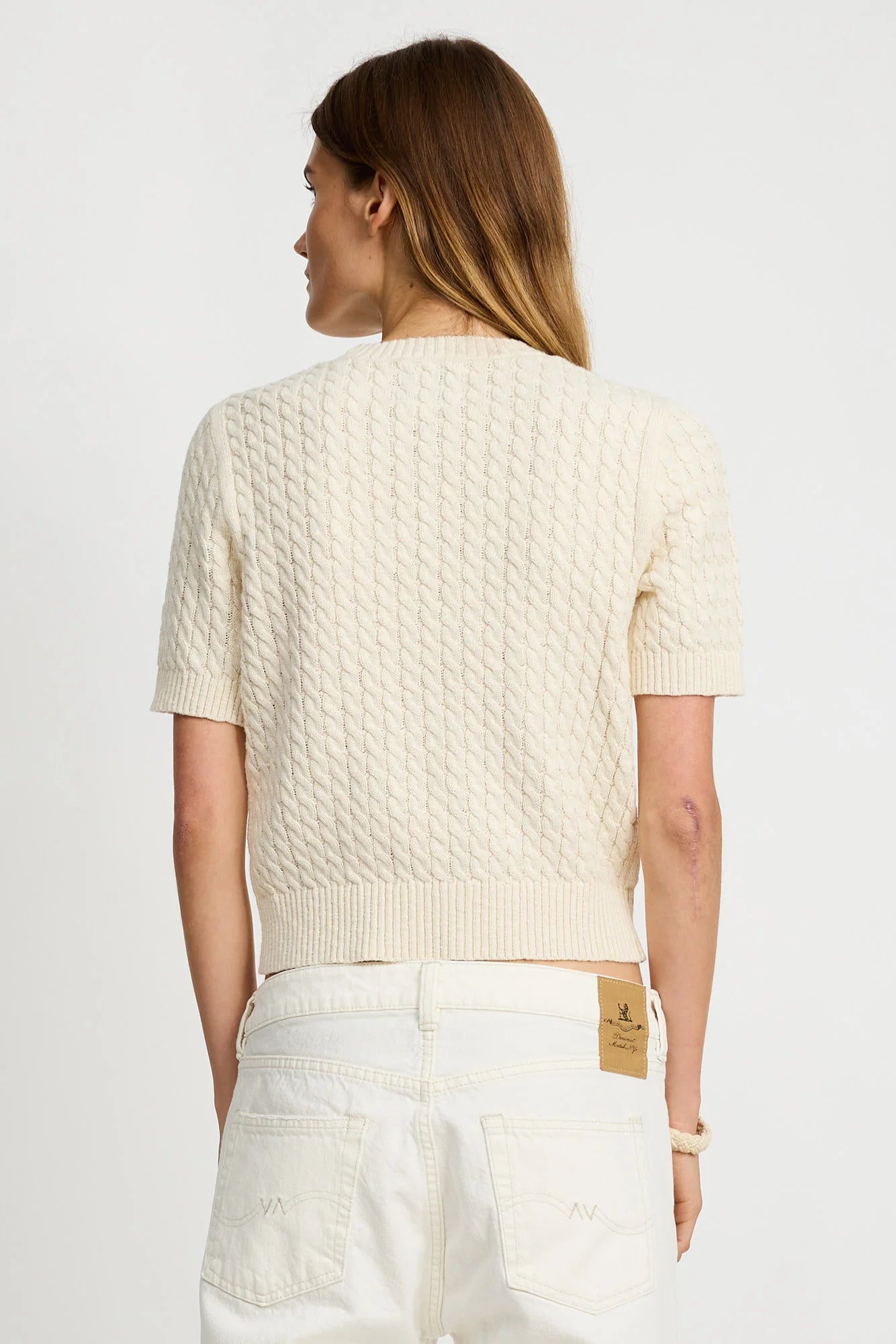 A person with long brown hair is seen from the back wearing the Denimist SHORT SLEEVE CABLE CREWNECK sweater in cream and high-waisted white jeans, set against a plain light background.