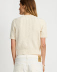 A person with long brown hair is seen from the back wearing the Denimist SHORT SLEEVE CABLE CREWNECK sweater in cream and high-waisted white jeans, set against a plain light background.