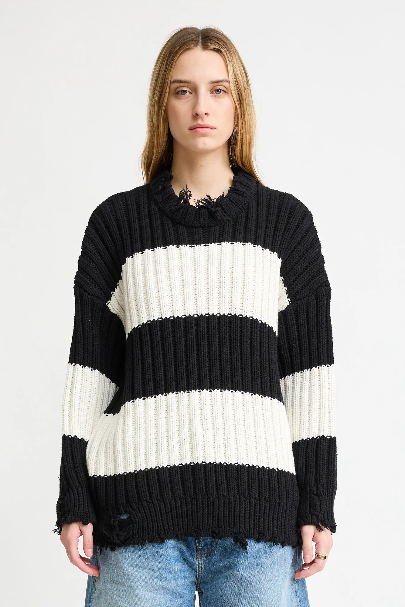 A person with long, straight hair models the Denimist OVERSIZED RIBBED SWEATER featuring black and white stripes and distressed edges, paired with blue jeans, against a plain white background.
