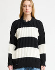 A person with long, straight hair models the Denimist OVERSIZED RIBBED SWEATER featuring black and white stripes and distressed edges, paired with blue jeans, against a plain white background.