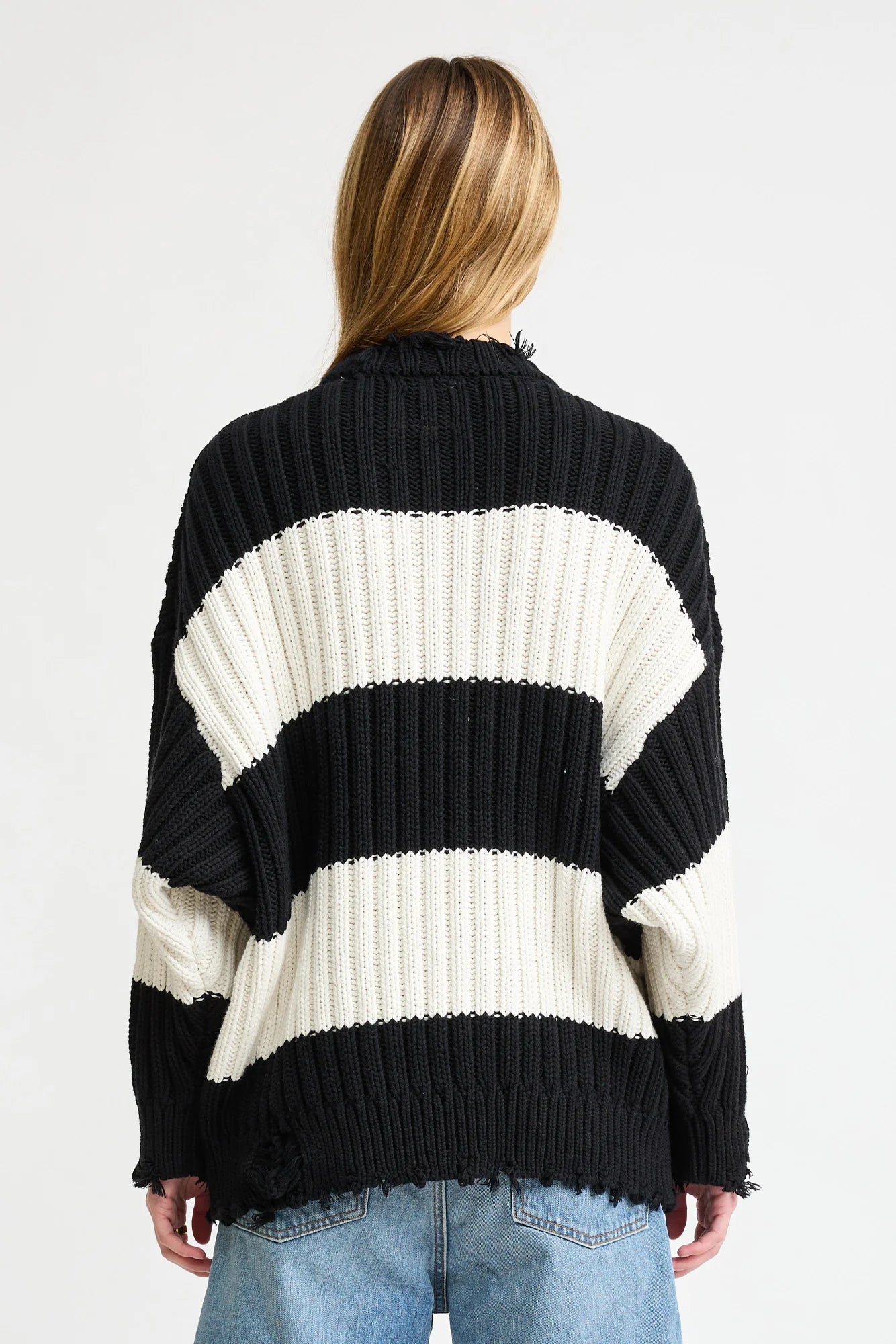 A person with long, light brown hair is seen from behind, wearing the Denimist OVERSIZED RIBBED SWEATER in black and white stripes and light blue jeans against a plain white background.