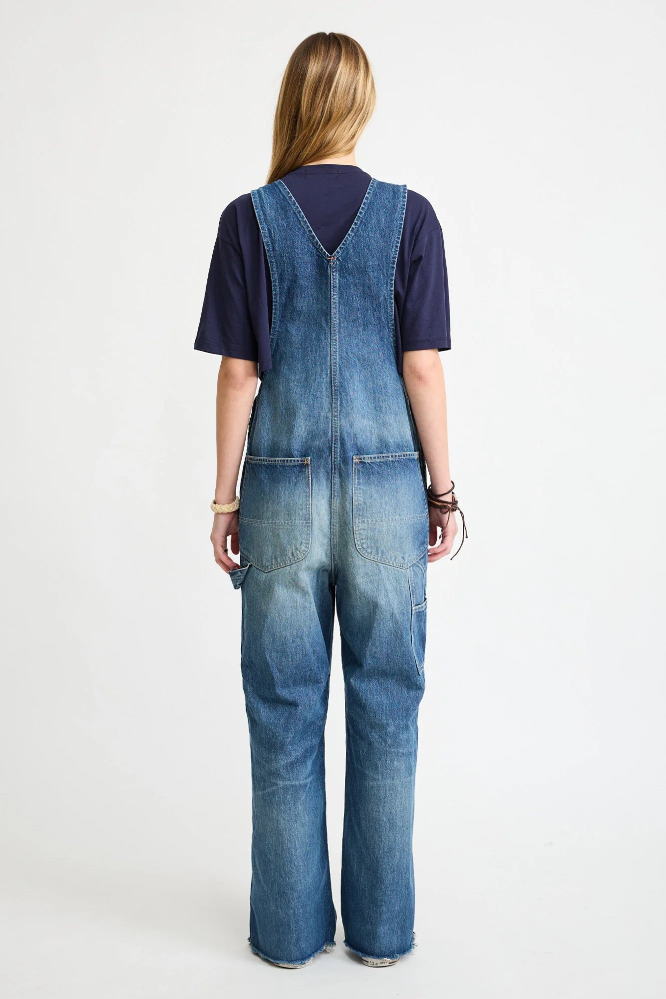 A person with long blonde hair faces away, wearing a dark T-shirt and Denimist’s SLOUCH BIB OVERALL in esher blue denim with back utility pockets. The background is plain white.