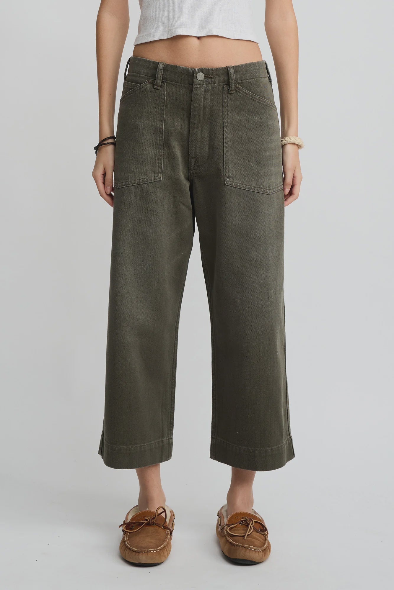 A person wearing Denimist’s Georgia Work Pant in olive green, featuring wide legs, 100% cotton fabric, and large front pockets, styled with a white cropped top and brown moccasin-style shoes against a plain light background.