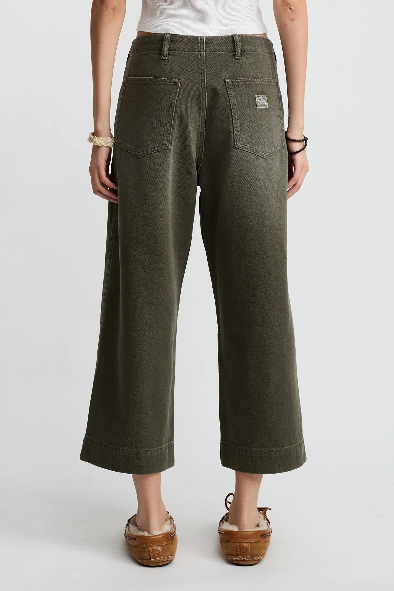 A person faces away, wearing Denimist's olive green 100% cotton Georgia Work Pant with a white top, brown moccasins, and bracelets on both wrists, set against a plain light background.