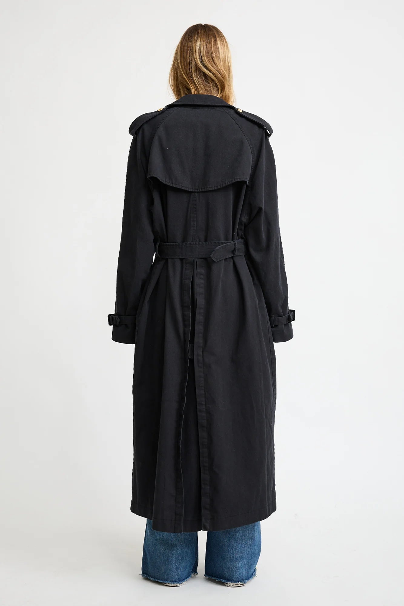 A person with long, light brown hair is seen from behind wearing the Denimist OVERSIZED TRENCH COAT with gold-tone buttons and wide lapels, paired with blue wide-leg jeans against a plain white background.