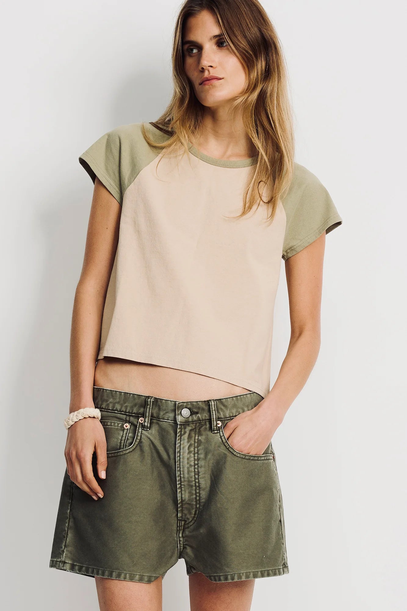 A woman with long, light brown hair stands with her hands in her pockets against a white background, wearing a beige and olive green short-sleeve top and Denimist HAISLEY SHORT relaxed-fit women's cotton shorts.
