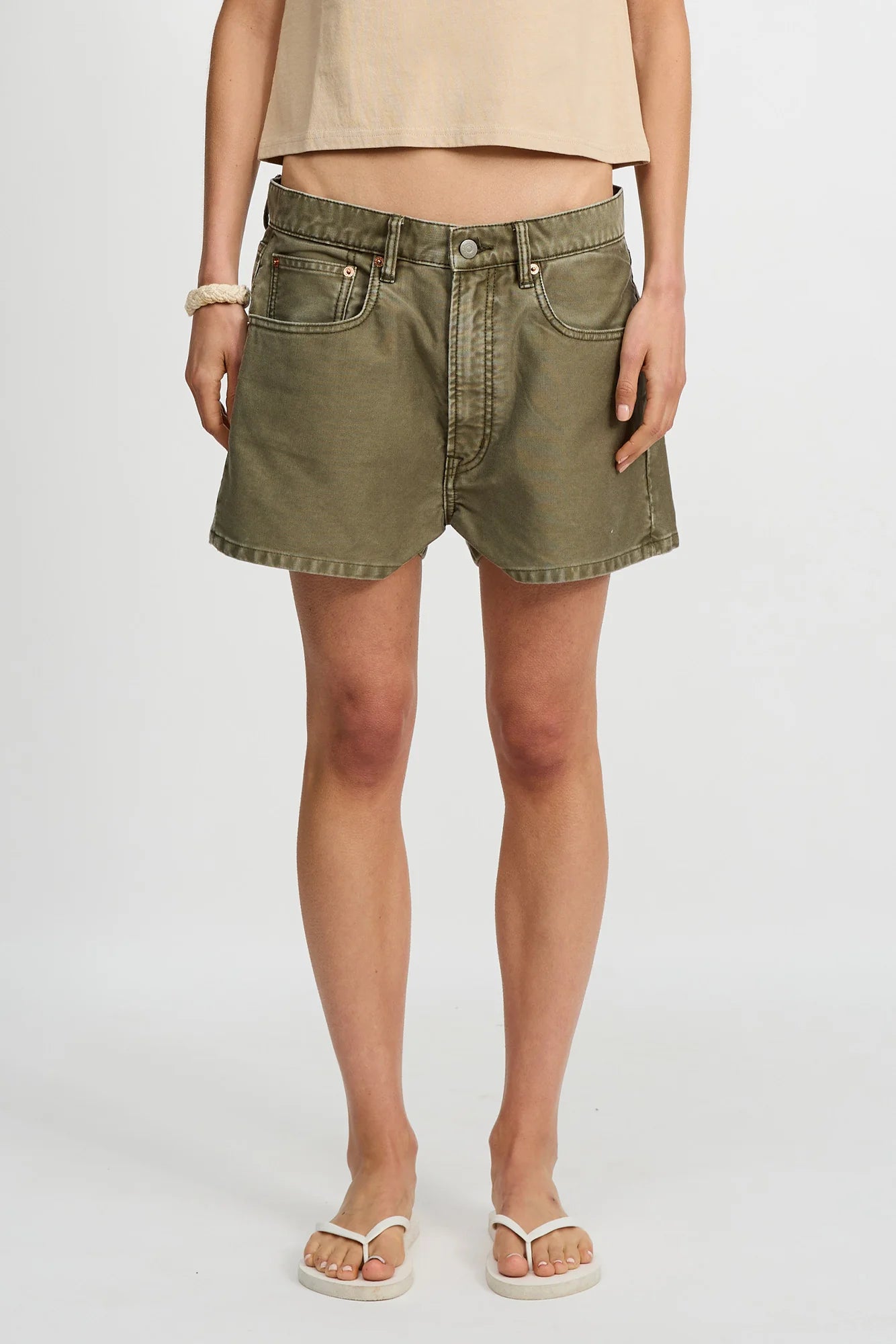 A person wears a beige top, Denimist HAISLEY SHORT relaxed-fit Bedford cord shorts in olive green, white flip-flops, and a light bracelet, standing against a plain white background.