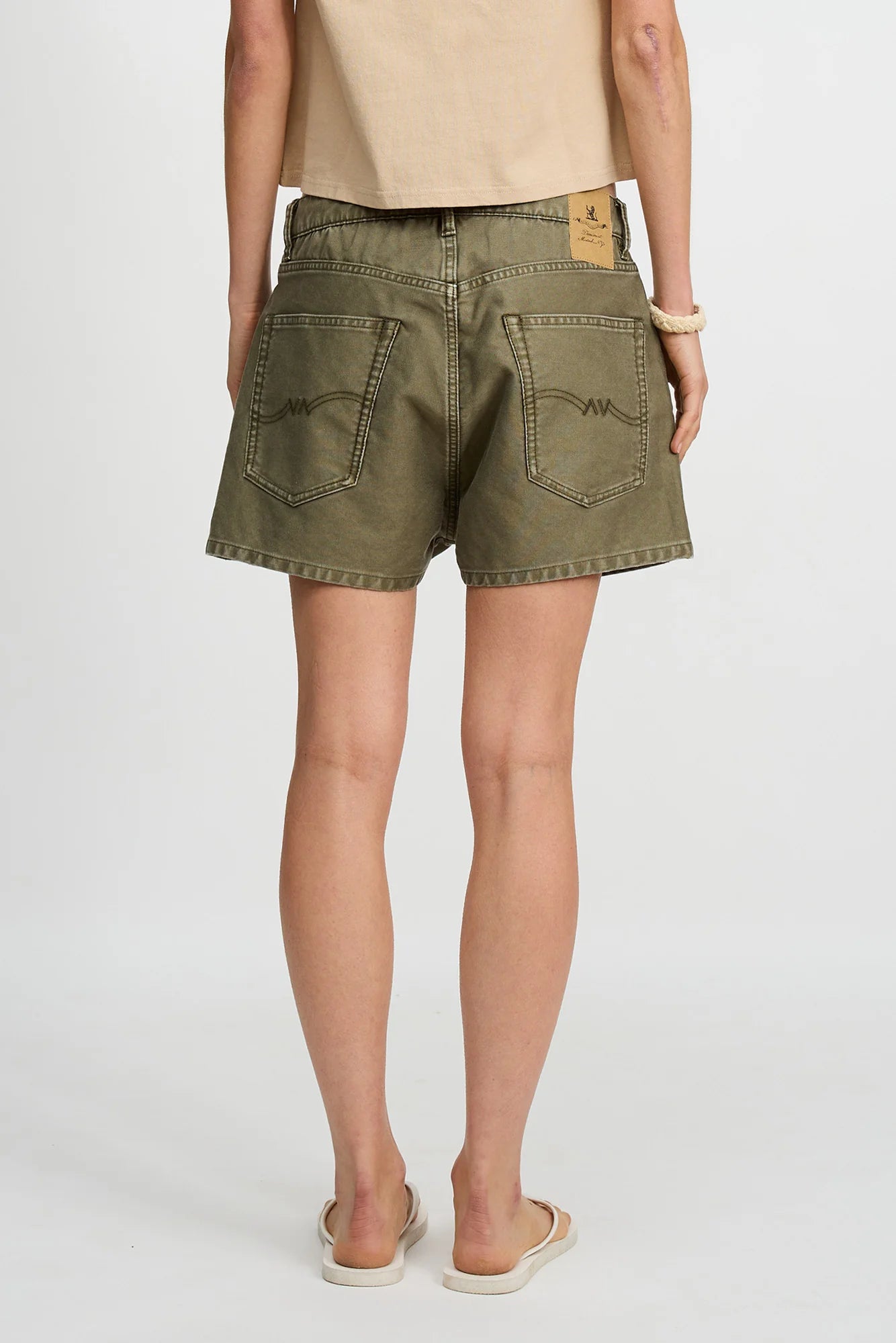 Model wears the Denimist HAISLEY SHORT in olive green, featuring large back pockets, paired with a tan tee, bracelet, and slip-on shoes. Shown from behind against a plain white background.