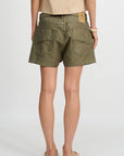 Model wears the Denimist HAISLEY SHORT in olive green, featuring large back pockets, paired with a tan tee, bracelet, and slip-on shoes. Shown from behind against a plain white background.