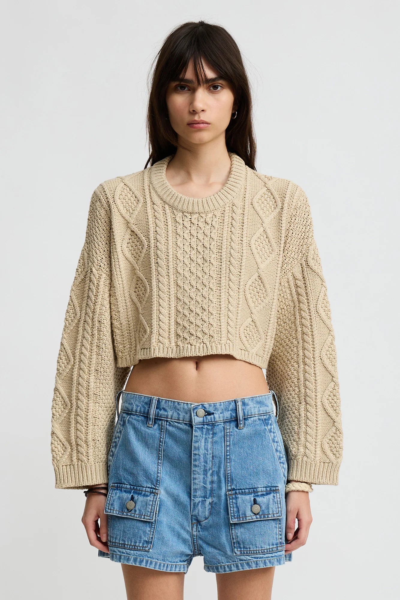 A woman with long brown hair wears a beige Denimist Cropped Cable Sweater and high-waisted blue denim shorts with front pockets, standing against a plain light background.