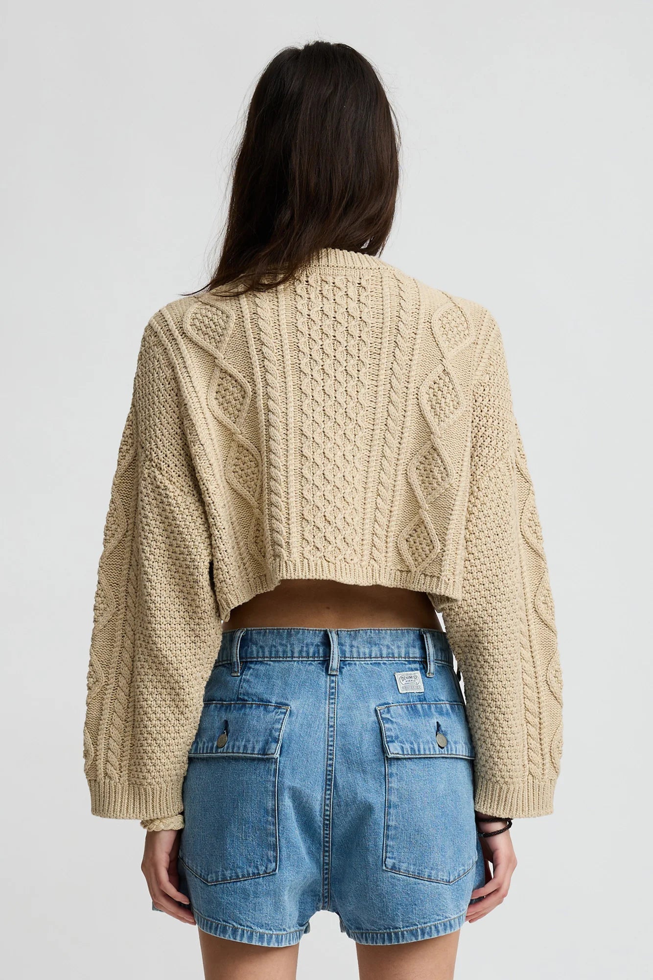 A person with long brown hair is seen from the back, wearing the Denimist Cropped Cable Sweater in beige and high-waisted blue denim shorts, standing against a plain light background.