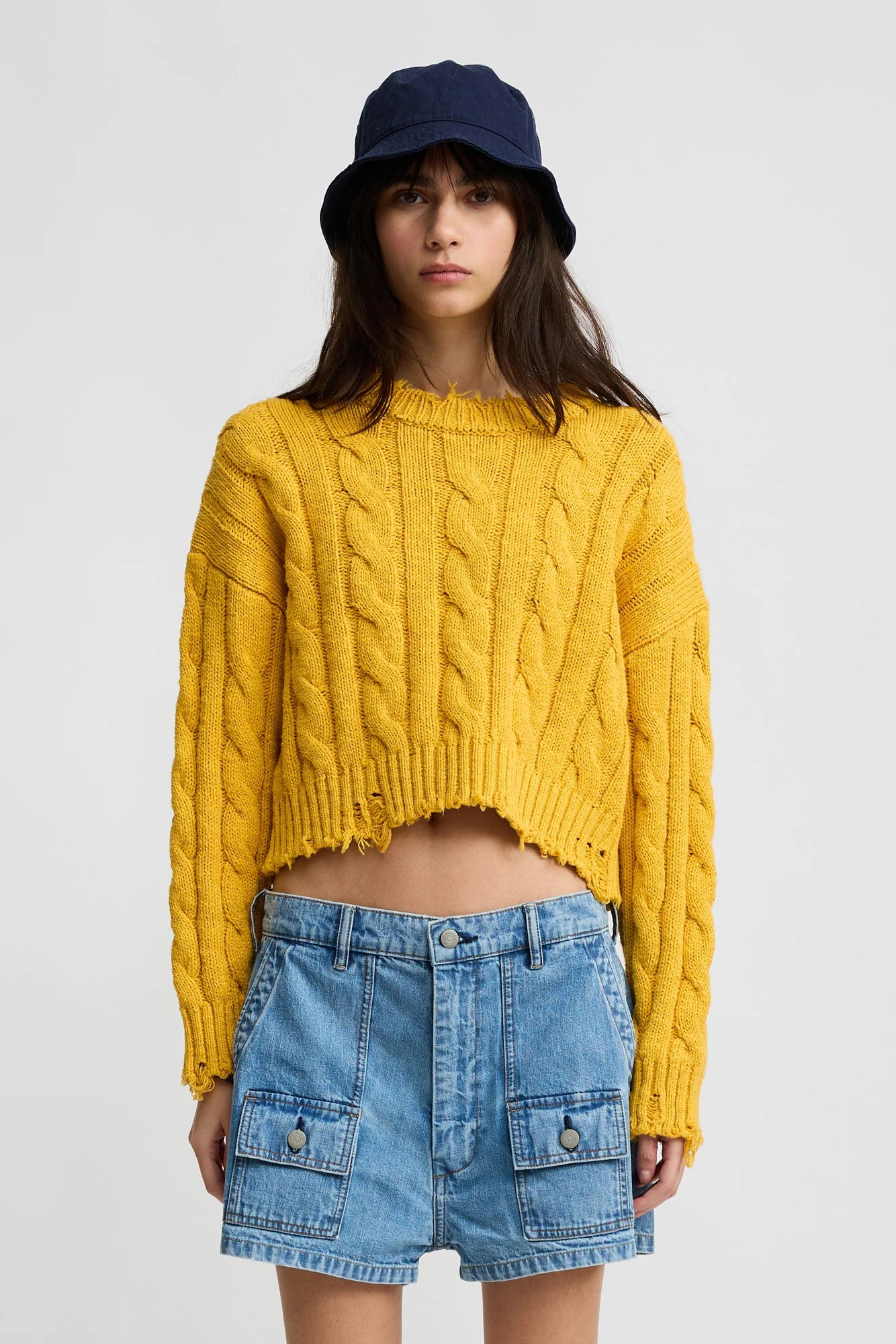 A woman stands against a plain light background wearing Denimist's Boxy Cable Sweater in yellow, paired with a navy bucket hat and high-waisted blue denim shorts with front pockets.