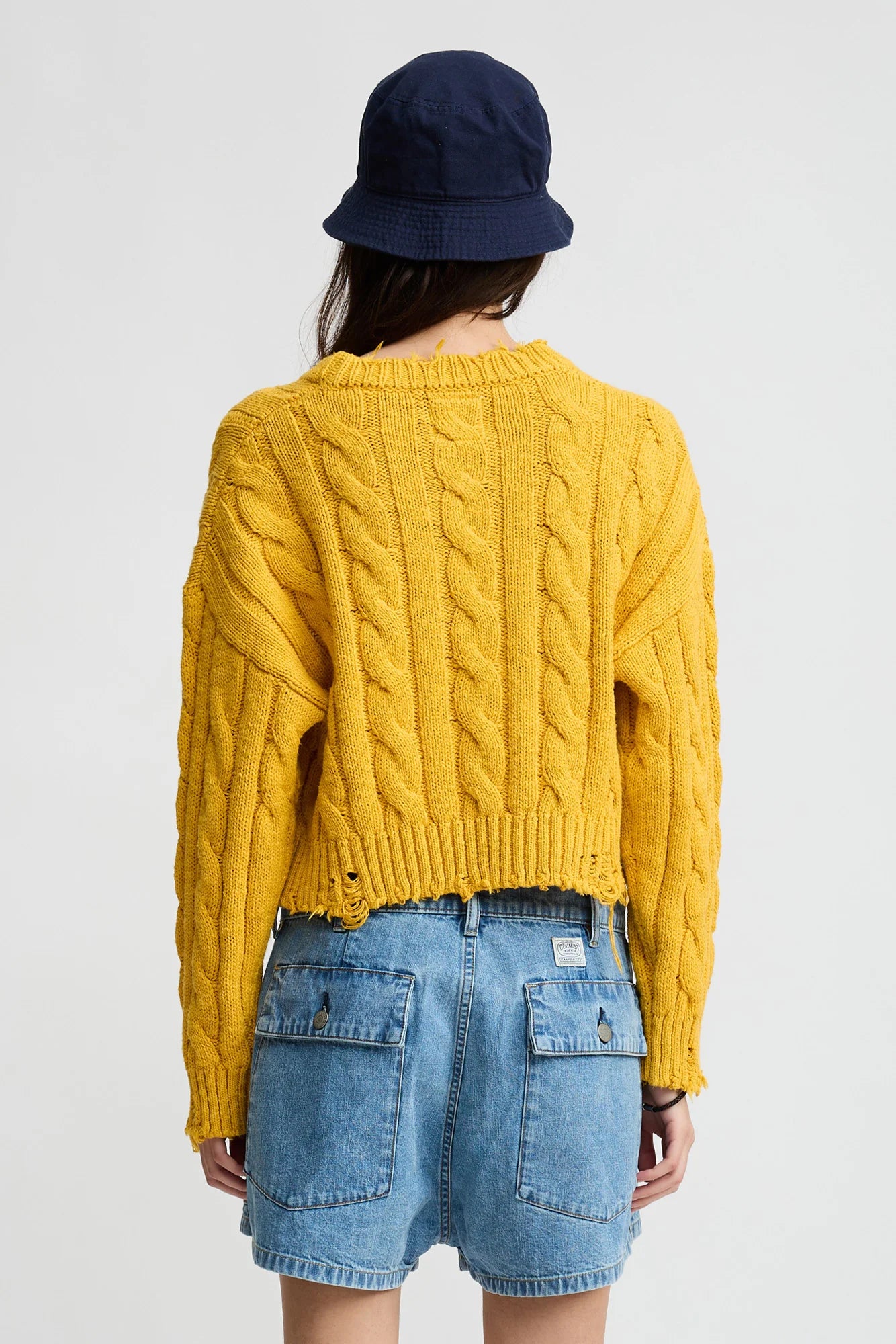A person wearing the Denimist Boxy Cable Sweater in yellow, a navy bucket hat, and a denim skirt with large back pockets stands facing away against a plain white background.