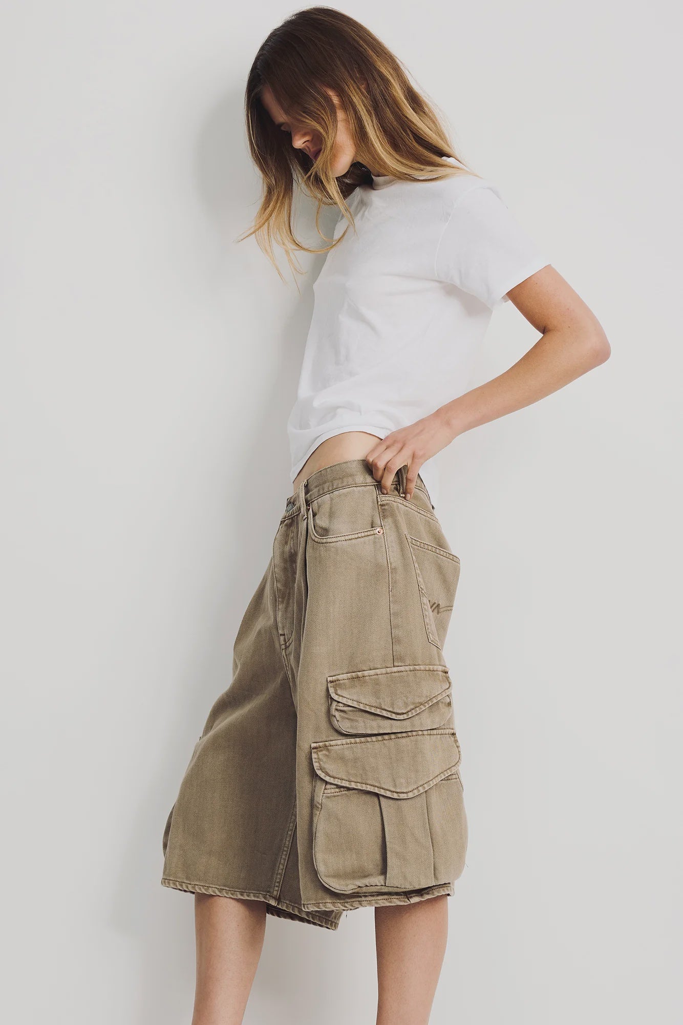 A woman stands against a white wall, wearing a plain white T-shirt and the Denimist KELBY CARGO SHORT—100% cotton, knee-length shorts with oversized flap pockets—looking down with her hand in her pocket.