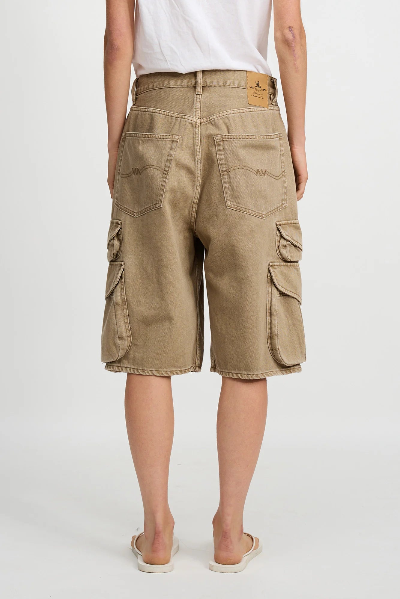 A person wearing a white t-shirt, Denimist KELBY CARGO SHORT in tan 100% cotton with oversized flap pockets, and beige slide sandals is shown from the back against a plain white background.