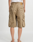 A person wearing a white t-shirt, Denimist KELBY CARGO SHORT in tan 100% cotton with oversized flap pockets, and beige slide sandals is shown from the back against a plain white background.