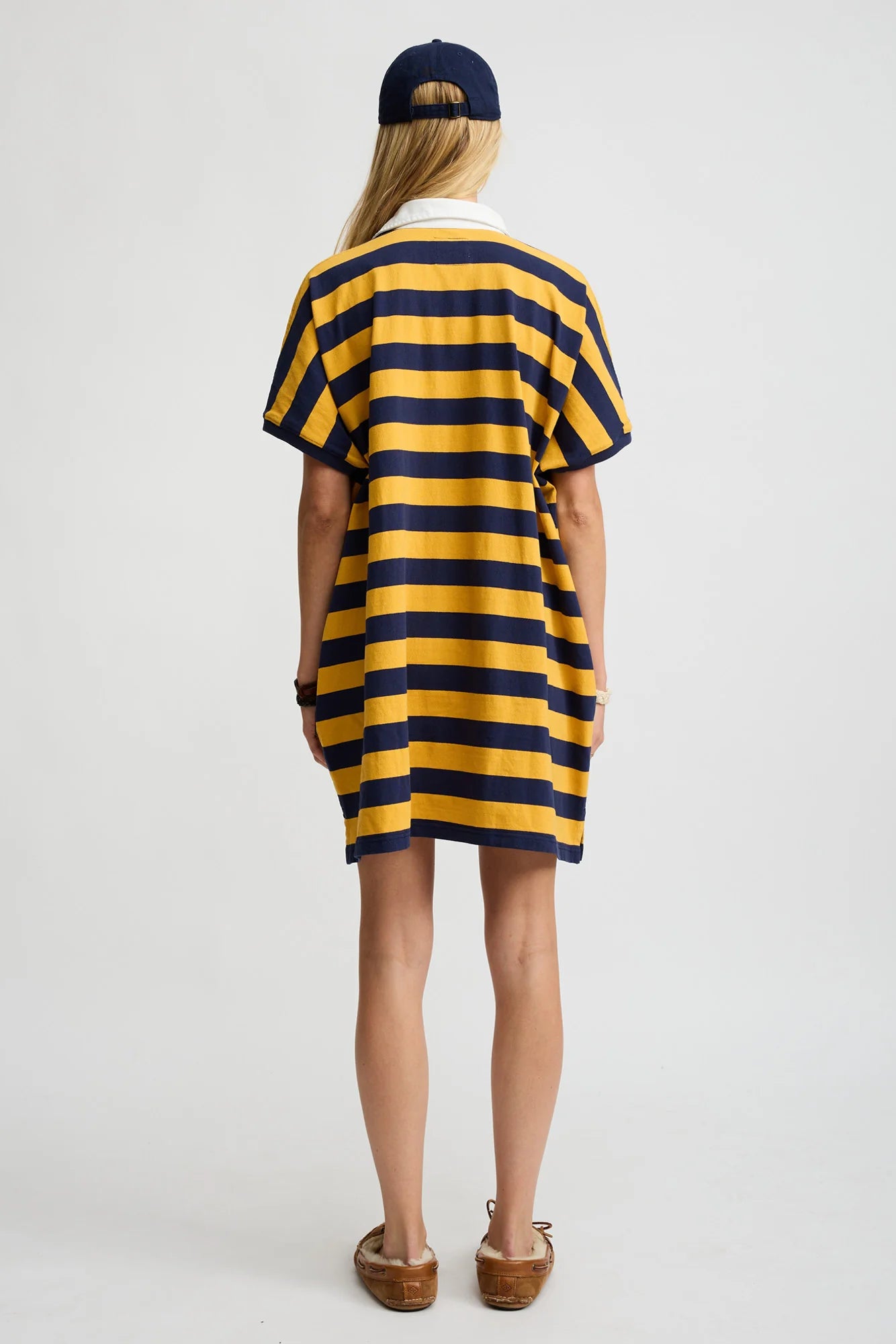 A person stands facing away, wearing the Denimist Rugby Shirt Dress—an oversized silhouette in navy and yellow stripes—paired with a navy cap and brown slip-on shoes, crafted from soft cotton, against a white background.