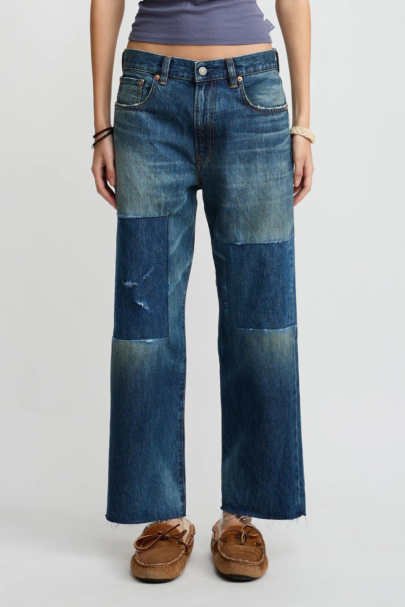 Someone stands against a plain light background wearing Denimist’s Lucy Boyfriend Jean—relaxed fit jeans with large, darker denim patches—paired with a purple tank top, moccasin-style shoes, and bracelets.