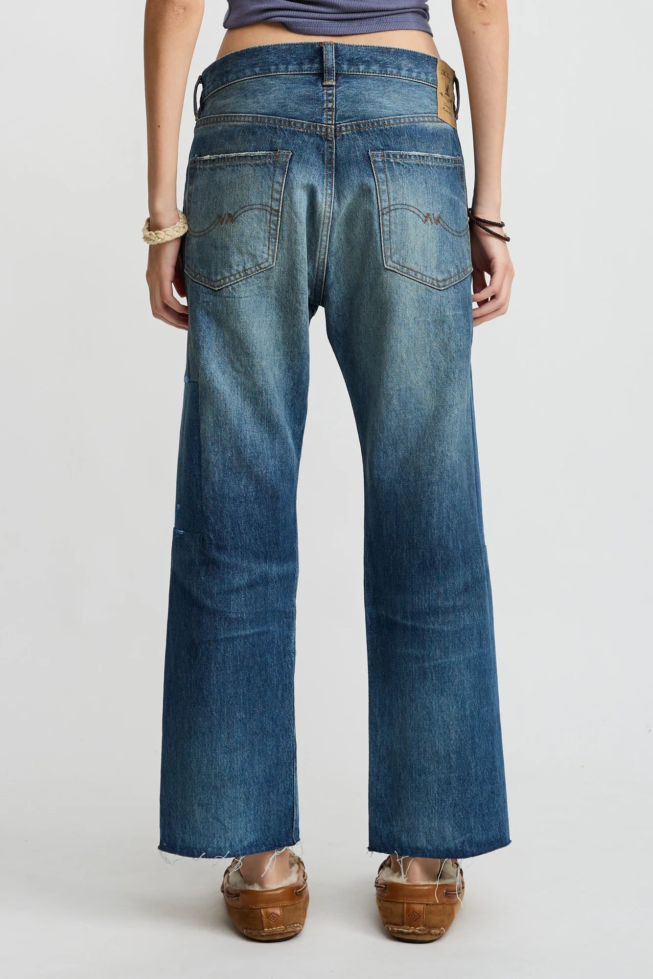 A person stands facing away, wearing Denimist's Lucy Boyfriend Jean with back pockets and a brown leather patch, a blue top, and brown platform sandals. Their hands rest by their sides, with bracelets on both wrists.