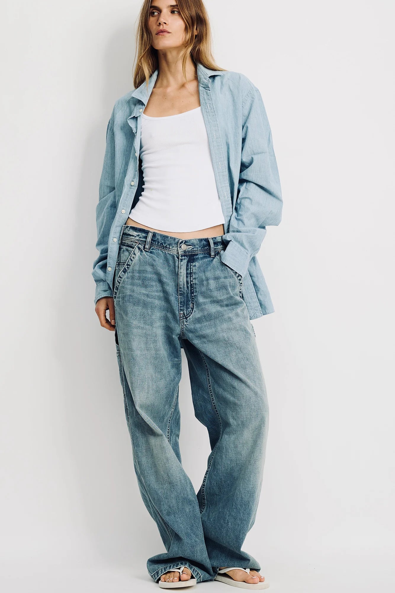 A woman stands against a plain background wearing the Denimist TERI WIDE LEG CARPENTER PANT, paired with a light blue denim shirt and white tank top. Her hands rest in her pockets, hair loose, highlighting relaxed style and soft cotton comfort.