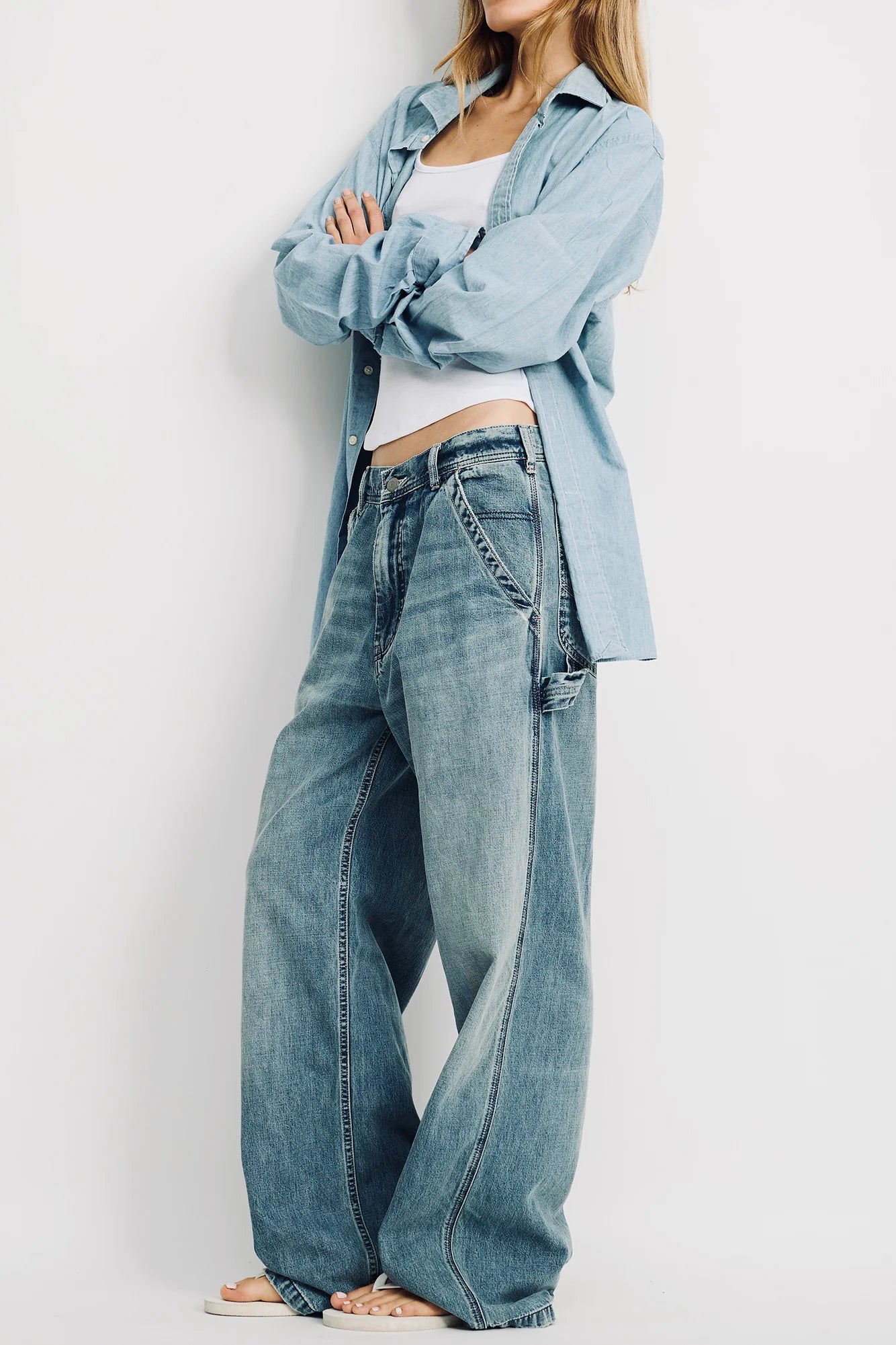 A woman stands with arms crossed, wearing a light blue shirt over a white top and Denimist's TERI WIDE LEG CARPENTER PANT. She is barefoot and facing slightly to the side against a plain white background.