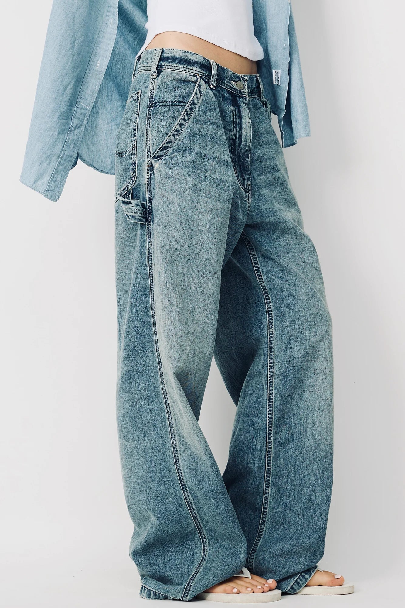 Wearing Denimist's TERI WIDE LEG CARPENTER PANT in blue, paired with a white top and unbuttoned light blue shirt. Photo shows the look from chest down, standing barefoot against a plain backdrop.