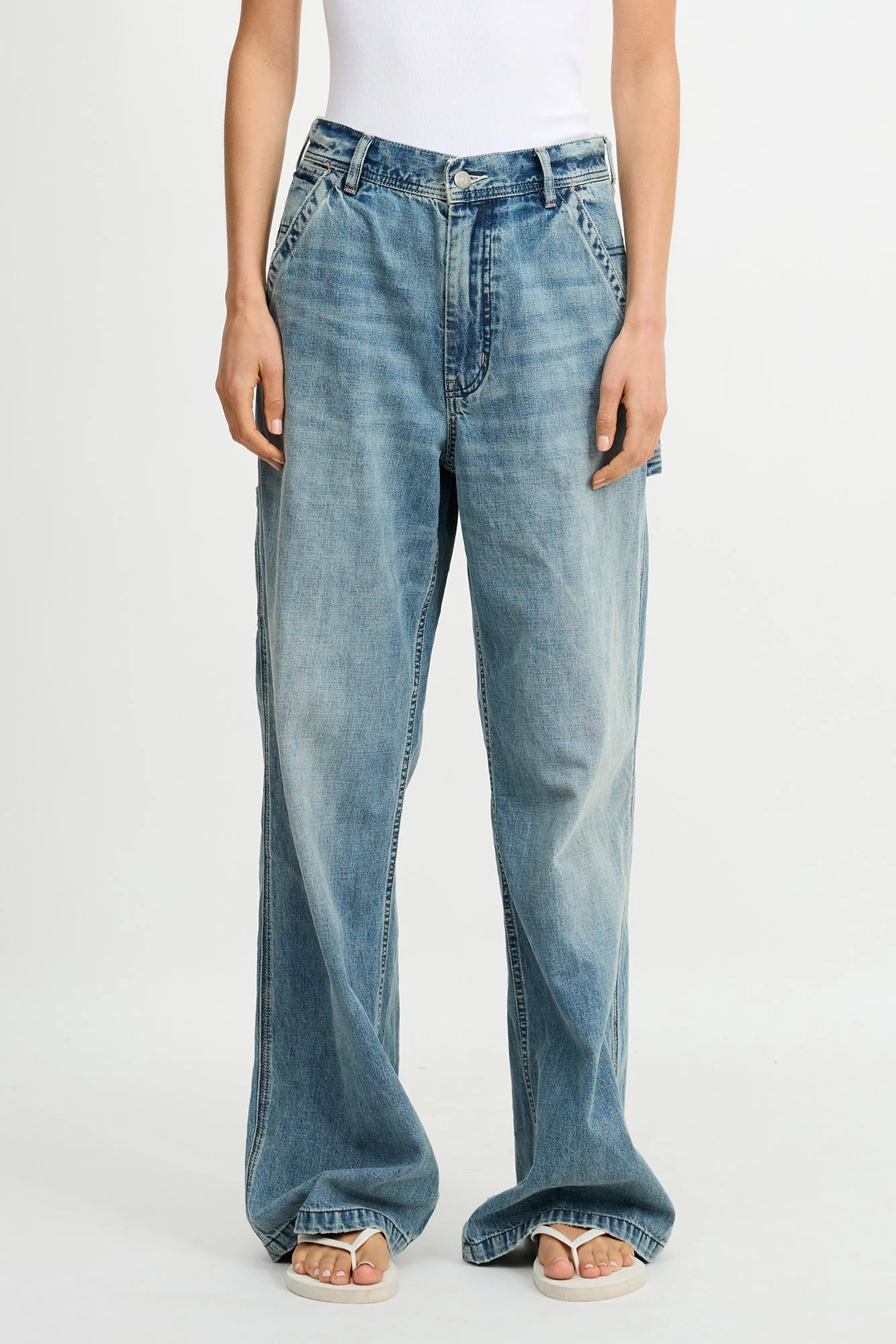 A person wears a white sleeveless top and Denimist TERI WIDE LEG CARPENTER PANT jeans with faded details, standing relaxed with open-toe sandals.