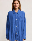 Wearing the Denimist SUTTON BUTTON FRONT BRIGHT BLUE oversized shirt with a front pocket and frayed denim shorts, a person with long hair stands against a plain light background.