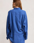 A woman with long brown hair stands with her back to the camera, wearing the Denimist SUTTON BUTTON FRONT BRIGHT BLUE shirt and white shorts against a plain light background.