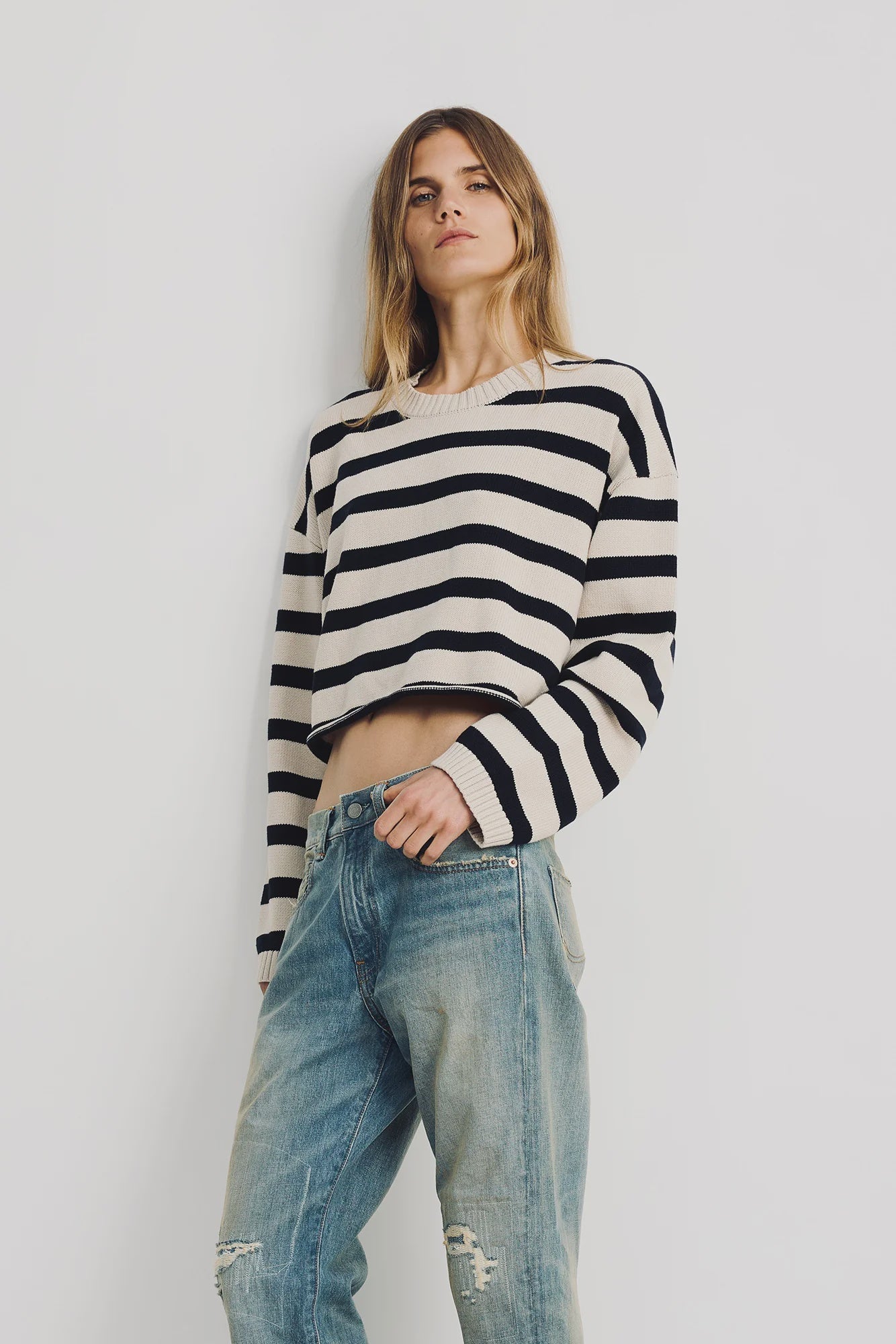 A person with long hair wears a Denimist CROPPED STRIPE SWEATER in black and white with loose, light blue ripped jeans, standing against a plain white background.