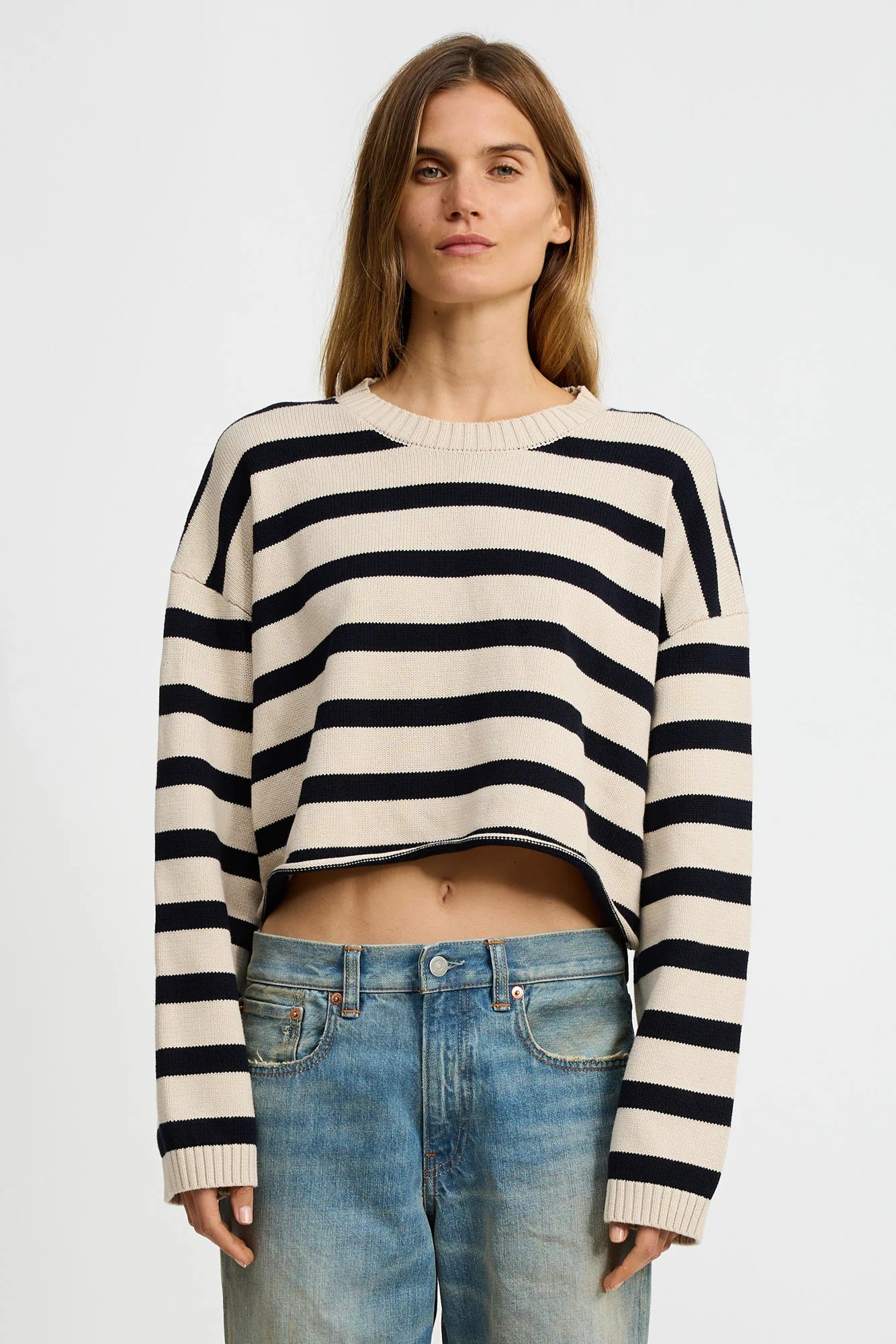 A person with long hair wears the Denimist CROPPED STRIPE SWEATER featuring black horizontal stripes, paired with blue jeans, and stands against a plain white background.