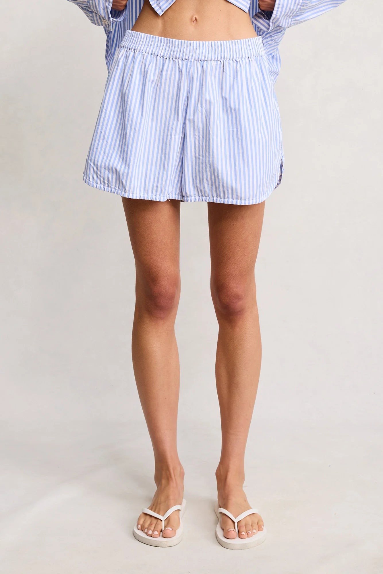 A person models Denimist PJ SHORTS, featuring light blue and white vertical stripes and an elastic waistband. Only their lower body is visible, paired with an open shirt and white sandals against a plain background.