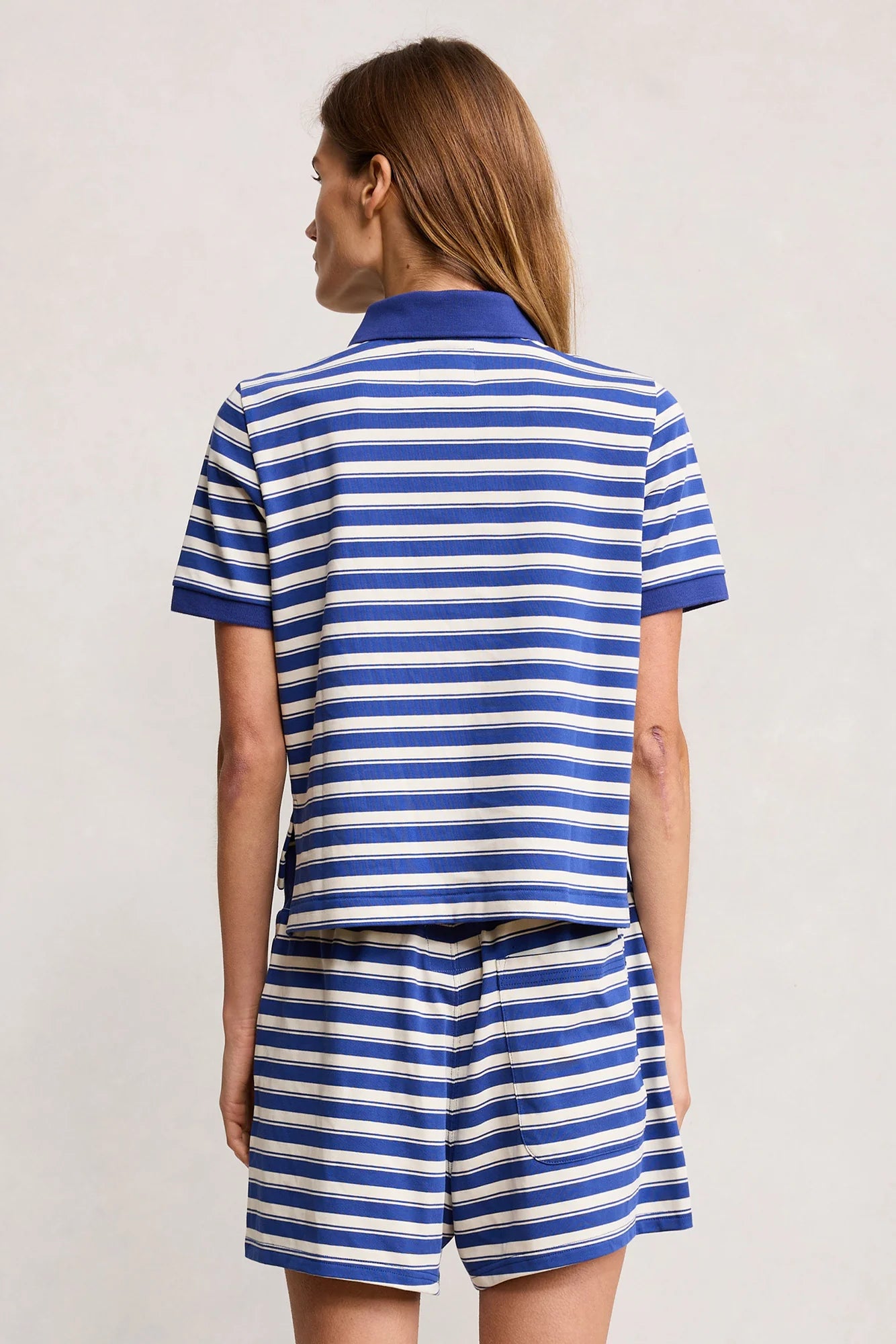 A person faces away, wearing Denimist's SHRUNKEN POLO—a blue and white striped cotton polo t-shirt and matching shrunken-fit shorts—against a plain light background.