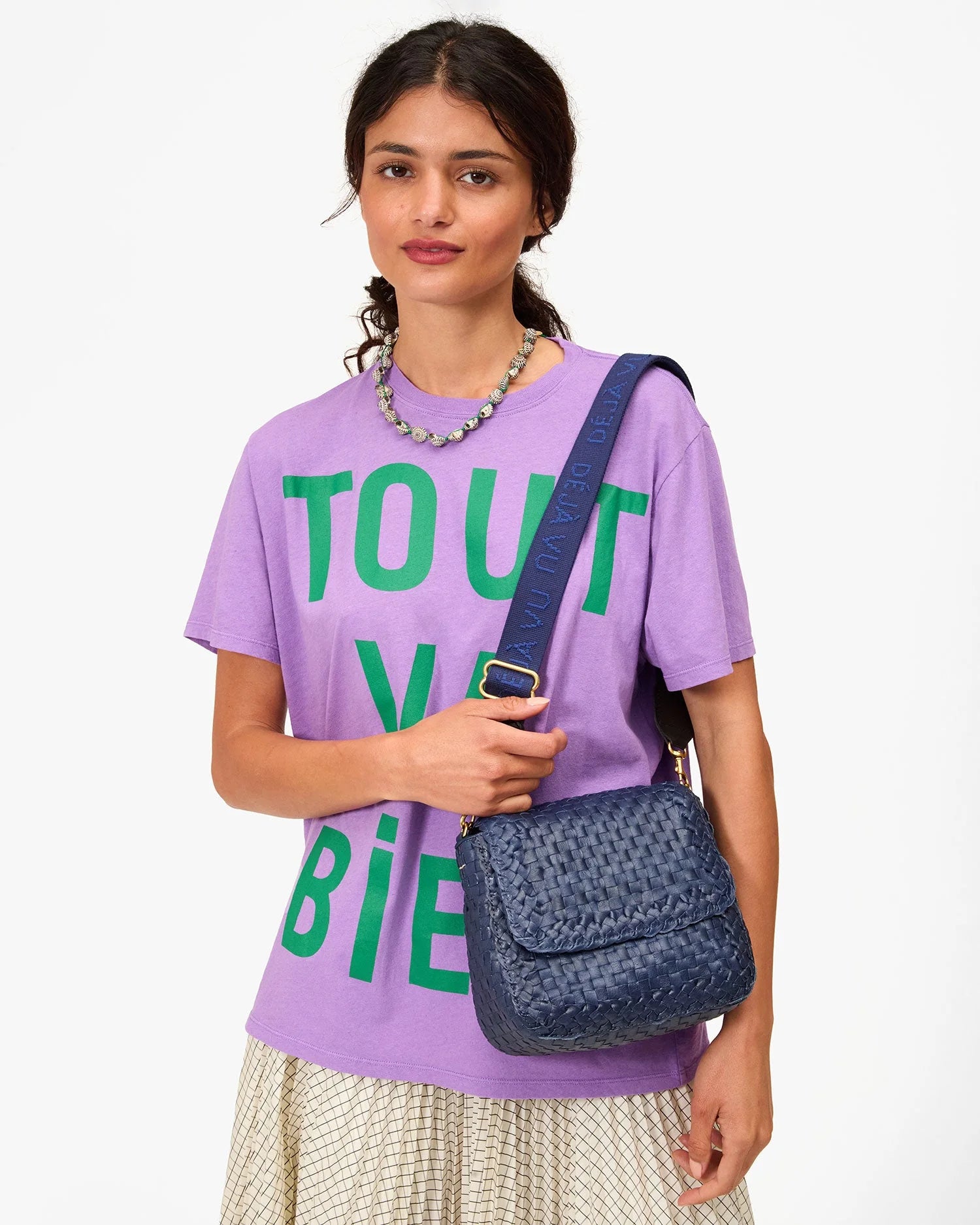 A woman in a lavender T-shirt, cream pleated skirt, and beaded necklace holds a blue bag with the Clare Vivier Adjustable Crossbody Strap, featuring cotton webbing, standing against a white background.