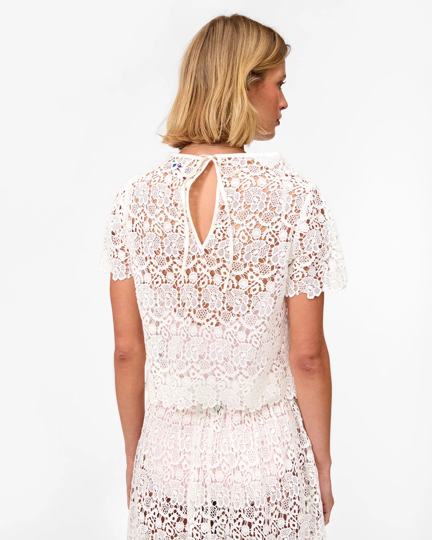 A woman with blonde hair is seen from behind wearing the Clare Vivier Lou Lou Top, a white, short-sleeve cotton lace blouse featuring a keyhole opening, neck tie closure, and sheer floral lace, paired with a matching skirt.