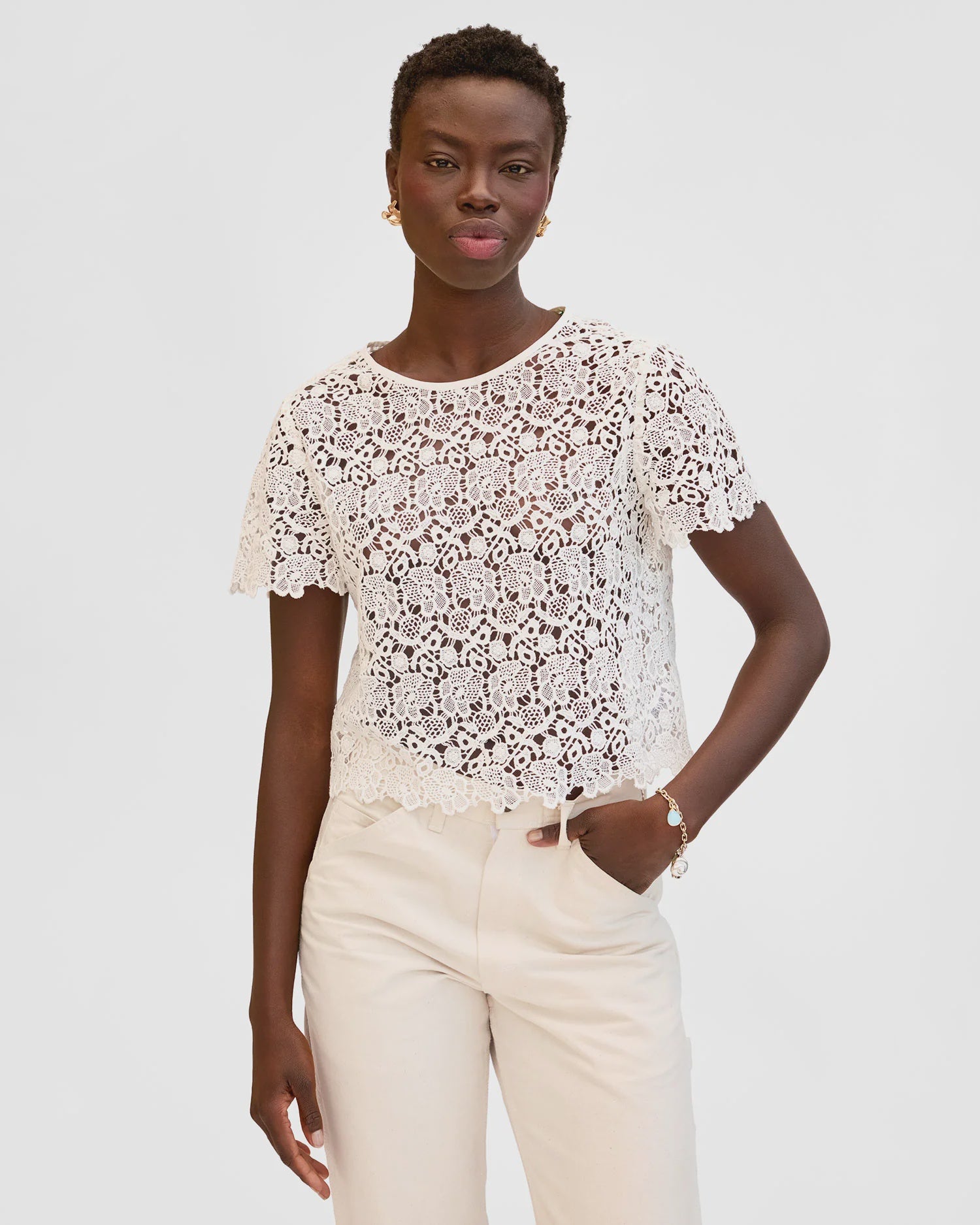 A woman with short hair stands against a plain light background, wearing the Clare Vivier Lou Lou Top—a white cotton lace short-sleeved blouse with a delicate neck tie—paired with light pants, gold earrings, and a bracelet.