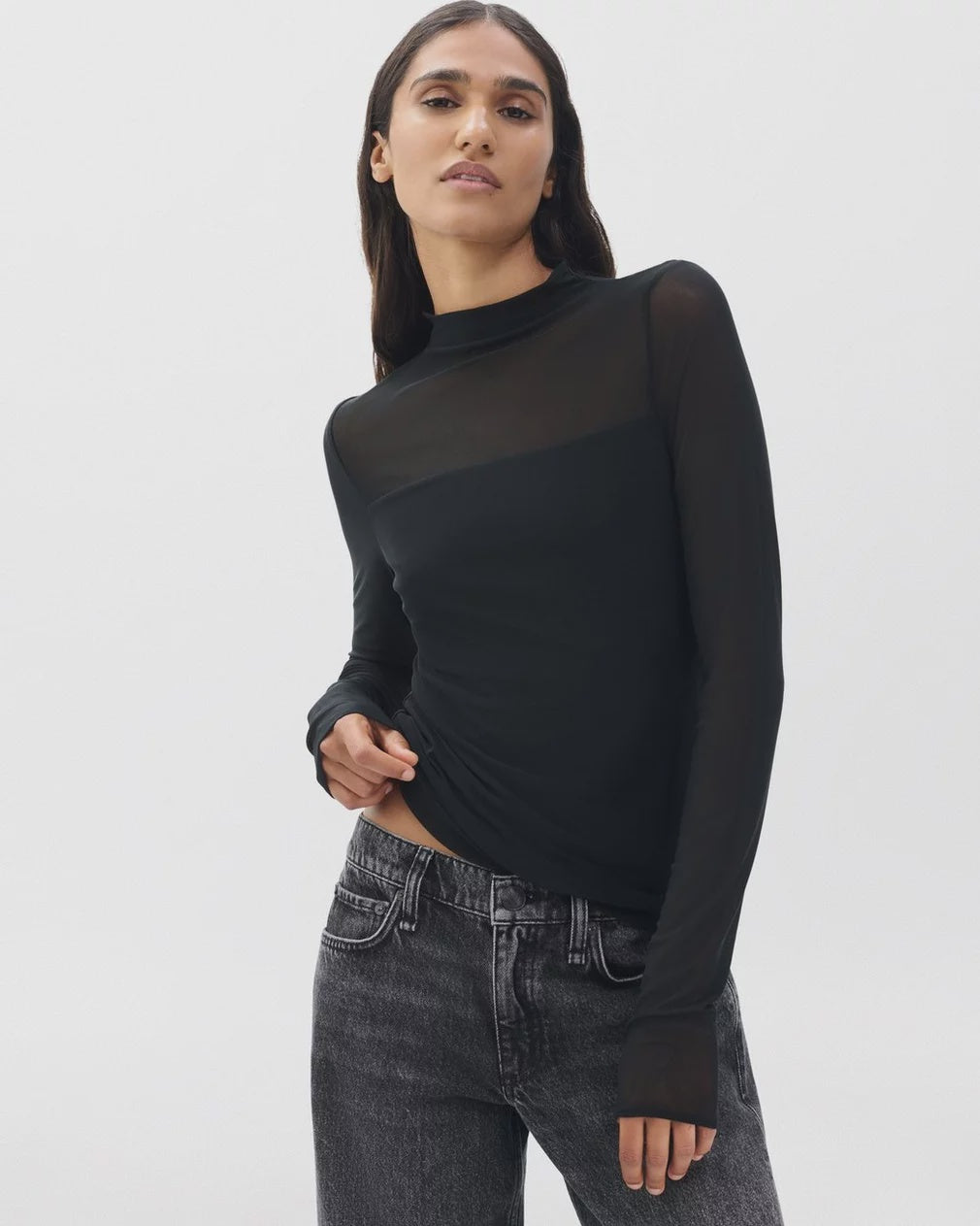 A woman with long brown hair wears the Rag & Bone luca dbl layer mix media mockneck ls top and faded black jeans, standing against a plain white background.