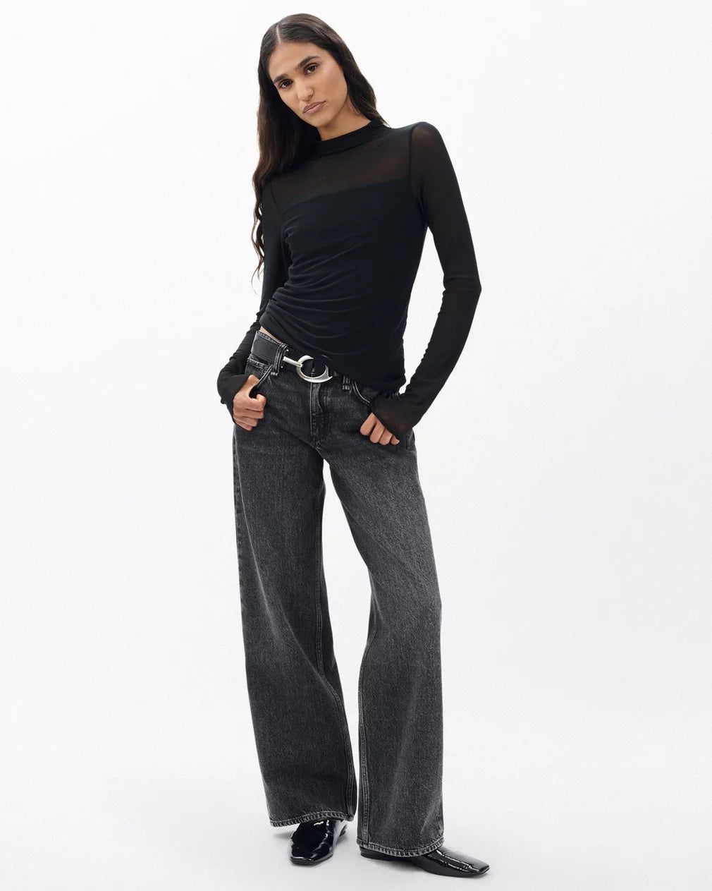 A woman poses confidently against a white background in the Rag & Bone luca dbl layer mix media mockneck long-sleeve top, styled with loose dark gray jeans and black pointed shoes. She has long wavy hair and a relaxed demeanor.
