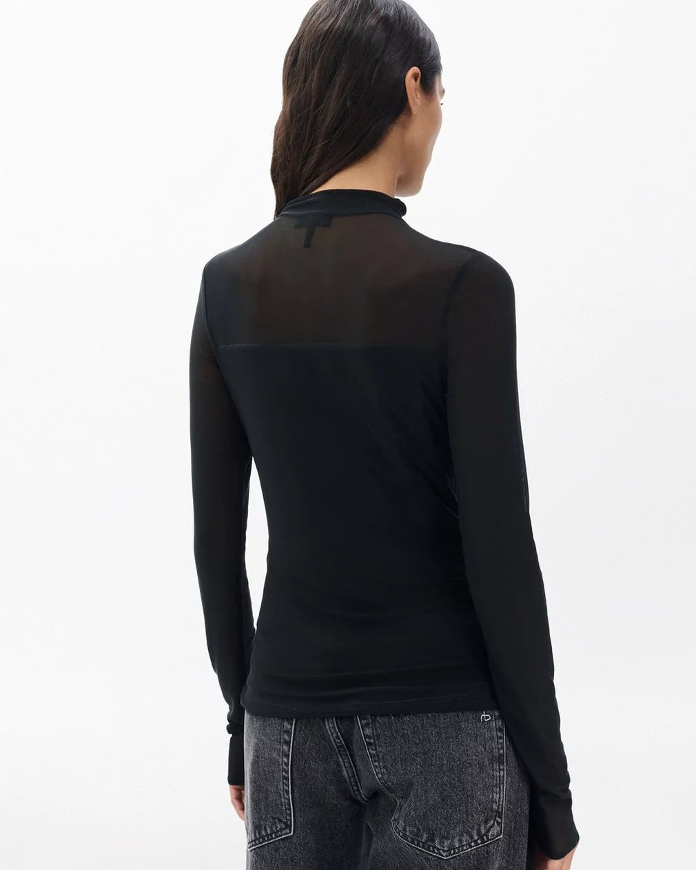A woman with long dark hair, seen from behind, wears the Rag & Bone “luca dbl layer mix media mockneck ls” semi-sheer black top and high-waisted black jeans against a plain white background.