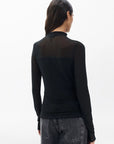 A woman with long dark hair, seen from behind, wears the Rag & Bone “luca dbl layer mix media mockneck ls” semi-sheer black top and high-waisted black jeans against a plain white background.