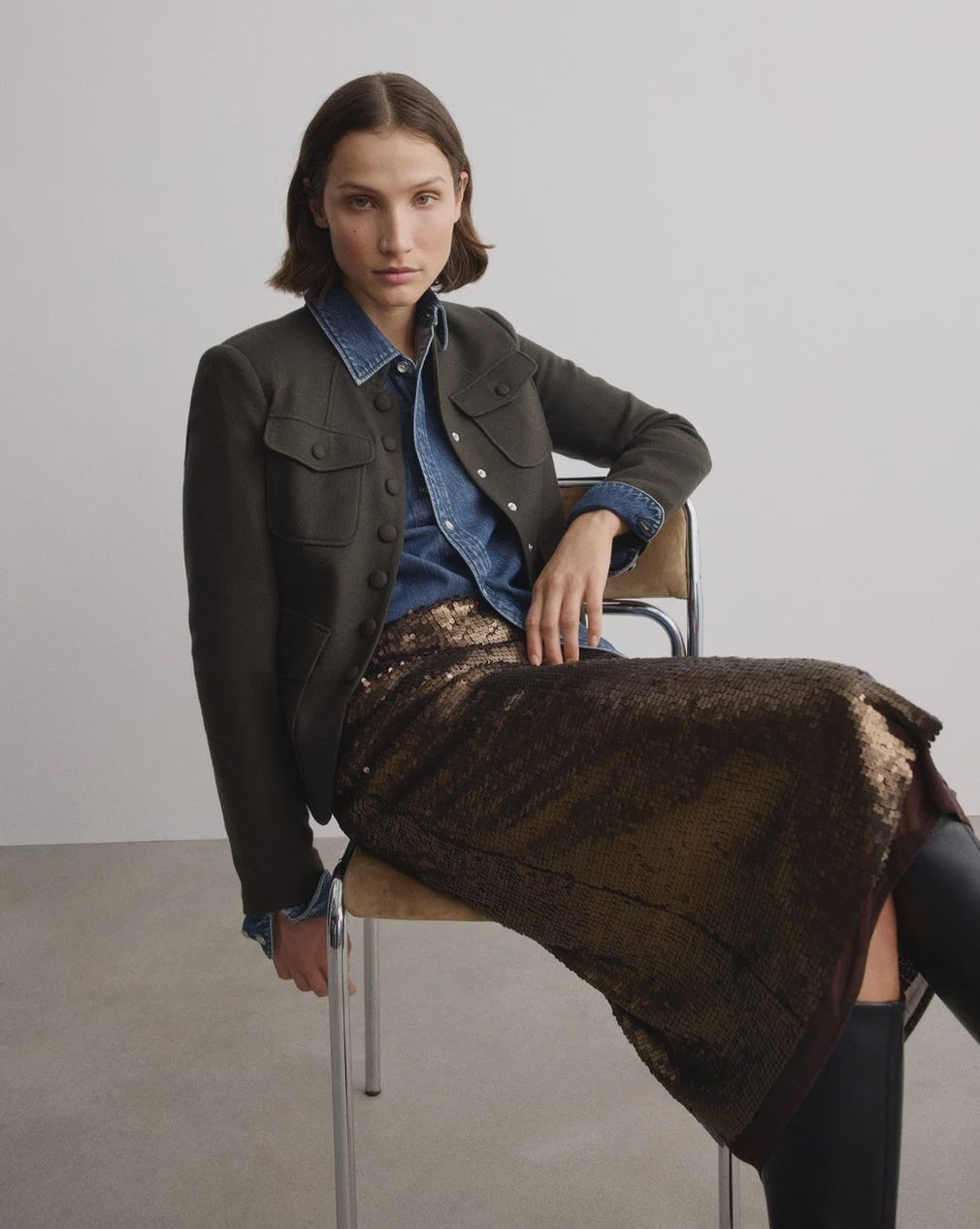 A woman with short hair sits indoors, wearing a dark jacket over a denim shirt, the Rag & Bone lucy sequin skirt, and black boots. She rests one arm on the chair’s back and has a neutral expression in this minimal setting.