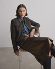 A woman with short hair sits indoors, wearing a dark jacket over a denim shirt, the Rag & Bone lucy sequin skirt, and black boots. She rests one arm on the chair’s back and has a neutral expression in this minimal setting.