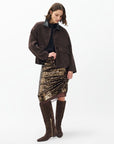 A woman stands against a white background in a dark oversized jacket, black top, Rag & Bone lucy sequin skirt, and knee-high brown suede boots. She looks down thoughtfully with one hand on her hip.